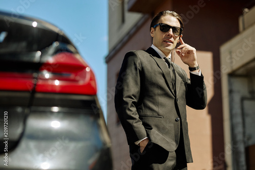 Portrait of young adult Caucasian businessman standing near car  speaking on earpiece and monitoring surroundings. Ideal for executive security  corporate transport  business travel