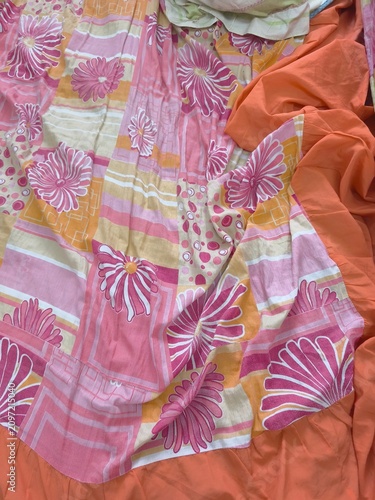 Close-up of patterned fabric featuring large stylized pink, white, and maroon flowers with geometric stripes in yellow, beige, and light pink, partly covered by orange cloth