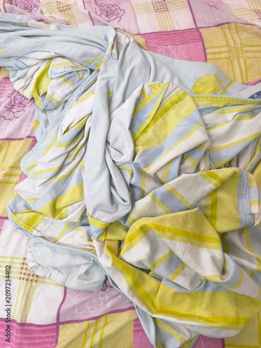 Close-up of bed showing colorful patchwork bedsheet with pink, purple, yellow floral squares and crumpled blue-yellow striped fabric loosely spread