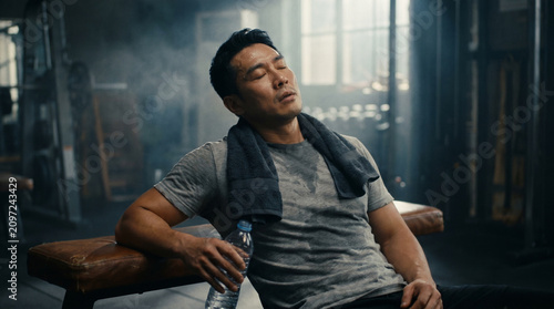 Exhausted Muscular East Asian Man Resting on Gym Bench After Intense Workout, Head Tilted Back with Eyes Closed Holding Water Bottle, Authentic Post-Workout Recovery and Relief in Moody Gym Lighting