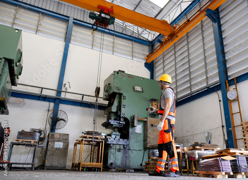 Industrial factory interior with a worker in safety gear inspecting a large hydraulic press beneath an overhead crane, surrounded by metal fabrication equipment and stacked materials.