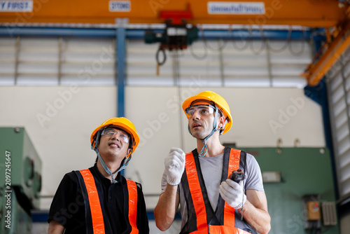 Two factory workers in hardhats operate an overhead crane inside an industrial workshop, wearing safety vests and gloves while inspecting equipment during lifting operations.