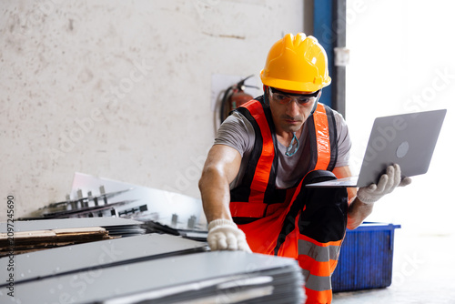 фотографія Construction worker in safety gear using a laptop to inspect stacked metal sheets in a warehouse, combining industrial labor with digital quality control
