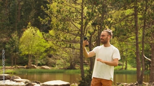 Man smiles while taking a selfie. He wears a white t-shirt and orange pants. Green trees and a river surround him. Sunlight filters through the forest canopy. Rocks line the water's edge