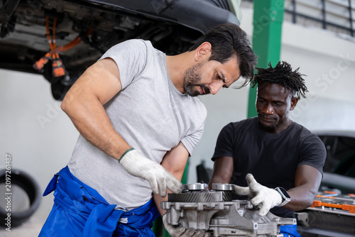 Two auto mechanics wearing gloves work together on a car transmission component in a professional garage, focusing on precise repair and maintenance beneath a lifted vehicle.