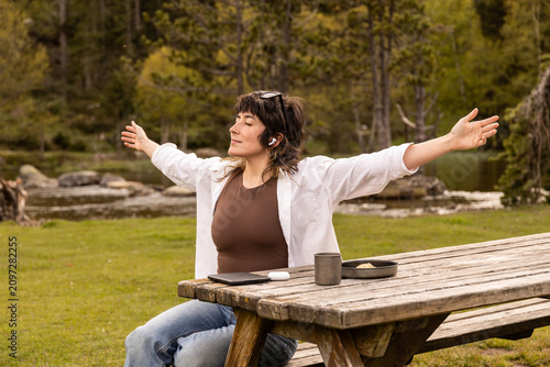 Woman with curly hair sits in a park. Her arm rests on a wooden railing. Sunglasses cover her eyes. Lush green grass and trees surround a small pond. Her calm expression shows peaceful thought