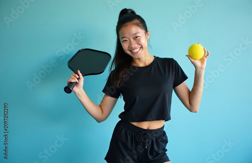 Young happy Asian woman holds black pickleball paddle, yellow ball. Person wears black sports clothes posing against light blue wall. Active girl smiling enjoys hobby recreation training practice