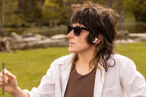 Woman with curly hair sits in a park. Her arm rests on a wooden railing. Sunglasses cover her eyes. Lush green grass and trees surround a small pond. Her calm expression shows peaceful thought