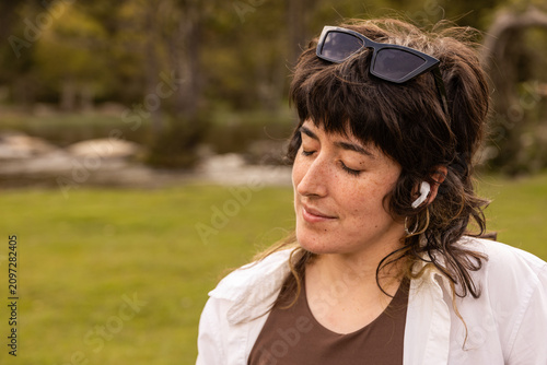 Woman with curly hair sits in a park. Her arm rests on a wooden railing. Sunglasses cover her eyes. Lush green grass and trees surround a small pond. Her calm expression shows peaceful thought