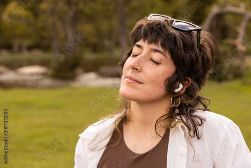 Woman with curly hair sits in a park. Her arm rests on a wooden railing. Sunglasses cover her eyes. Lush green grass and trees surround a small pond. Her calm expression shows peaceful thought