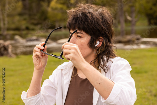 Woman with curly hair sits in a park. Her arm rests on a wooden railing. Sunglasses cover her eyes. Lush green grass and trees surround a small pond. Her calm expression shows peaceful thought