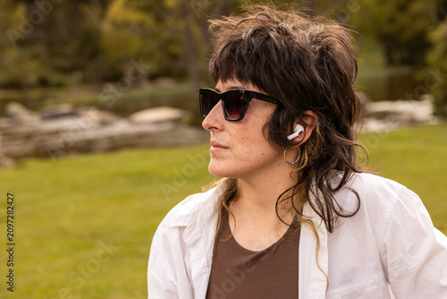 Woman with curly hair sits in a park. Her arm rests on a wooden railing. Sunglasses cover her eyes. Lush green grass and trees surround a small pond. Her calm expression shows peaceful thought