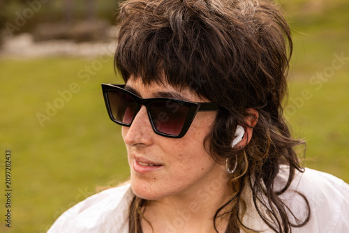 Woman with curly hair sits in a park. Her arm rests on a wooden railing. Sunglasses cover her eyes. Lush green grass and trees surround a small pond. Her calm expression shows peaceful thought