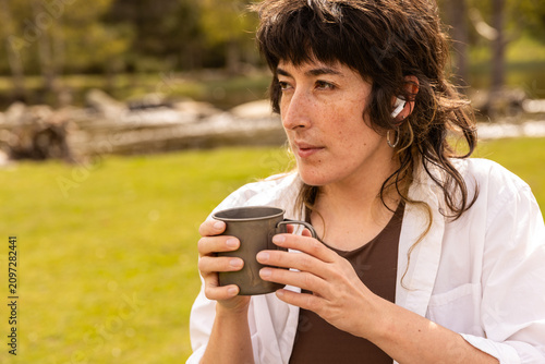 Woman sits at a wooden picnic table in a green park. She holds a coffee cup, with a tablet on the table. Her calm expression shows contentment. Ideal for work breaks or outdoor leisure scenes