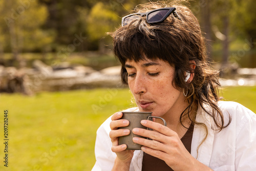 Woman sits at a wooden picnic table in a green park. She holds a coffee cup, with a tablet on the table. Her calm expression shows contentment. Ideal for work breaks or outdoor leisure scenes