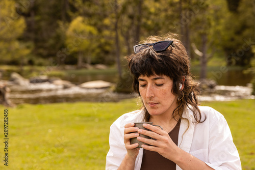 Woman sits at a wooden picnic table in a green park. She holds a coffee cup, with a tablet on the table. Her calm expression shows contentment. Ideal for work breaks or outdoor leisure scenes