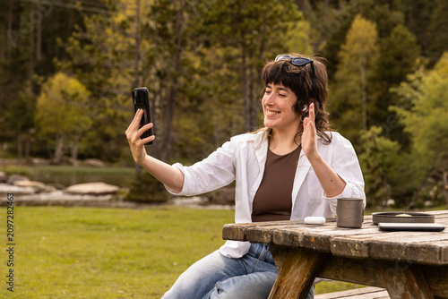 Woman smiles while taking a selfie at a wooden picnic table. She wears a white cardigan over a brown top. Her arms stretch wide as she captures the moment. Coffee mug and book rest on the bench