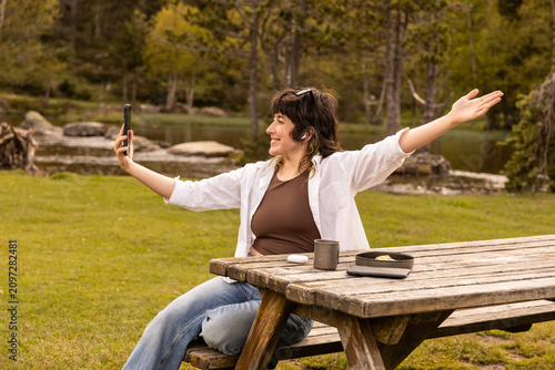 Woman smiles while taking a selfie at a wooden picnic table. She wears a white cardigan over a brown top. Her arms stretch wide as she captures the moment. Coffee mug and book rest on the bench