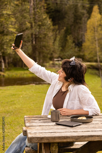 Woman smiles while taking a selfie at a wooden picnic table. She wears a white cardigan over a brown top. Her arms stretch wide as she captures the moment. Coffee mug and book rest on the bench