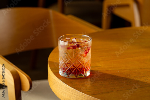 A drink with ice and fruits rests on a wooden table at a cozy cafe. The light from the afternoon sun highlights the glass and the colors inside it