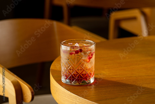 A drink sits on a wooden table in a cafe. The glass contains ice and red fruit. Soft light illuminates the scene. Chairs are placed around the table