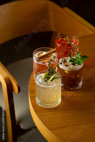 Four cocktails sit on a wooden table in a relaxed space. Each drink has unique ingredients and garnishes, showing a variety of colors. People may be enjoying time together