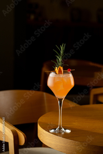 A drink sits on a wooden table in a bar. The cocktail has orange slices, cherries, and a sprig of herb on top. The light is low, creating a cozy atmosphere in the evening