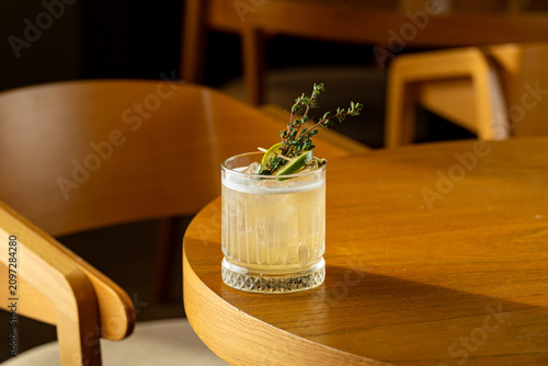 A clear drink sits in a glass on a wooden table. The drink has a garnish of herbs and a slice of lemon. Sunlight brightens the lounge area