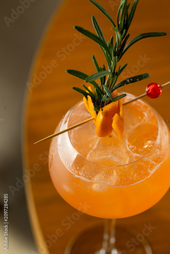 A glass filled with a bright drink sits on a wooden table. The drink is garnished with an orange twist and a sprig of rosemary. It is evening in a bar setting