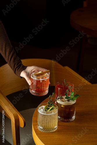 A person holds a cocktail while sitting at a wooden table in a bar. Three other drinks with garnishes sit on the table. The atmosphere is relaxed and warm in the evening light