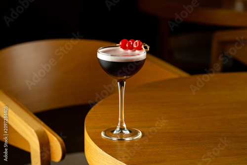 A cocktail glass filled with dark liquid is placed on a wooden table. Red cherries are garnished on top. The setting is warm and intimate, suitable for social gatherings in the evening