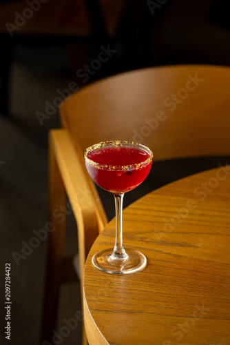 A cocktail with a bright red liquid sits in a delicate glass on a wooden table. The setting is cozy, with soft lighting enhancing the scene