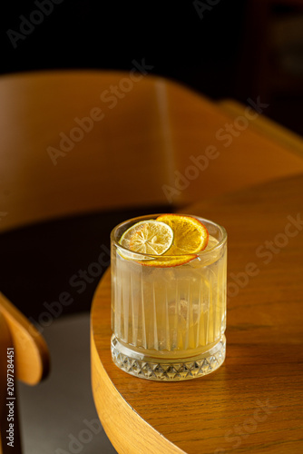 A clear drink with dried orange and lemon slices rests on a wooden table. The setting is a cafe during the evening hours. Soft lighting creates a warm atmosphere
