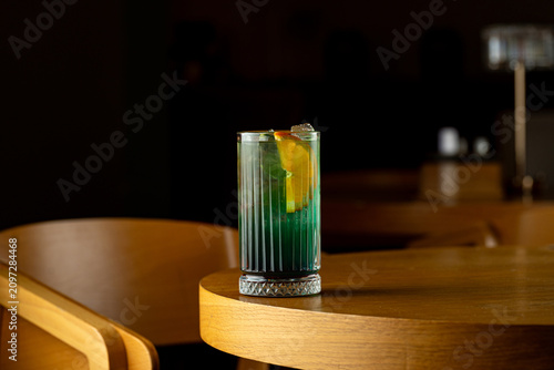 A drink placed on a wooden table shows layers of green and orange with ice. The setting is a dimly lit bar during evening hours