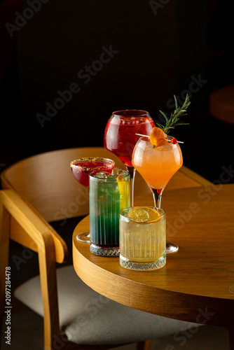 Brightly colored drinks are placed on a wooden table at a bar. People are enjoying the night, sipping different cocktails. The atmosphere is lively and relaxed