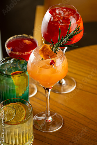 A group of mixed drinks is displayed on a wooden table. Each drink has different colors and garnishes. The setting is at a bar during the evening
