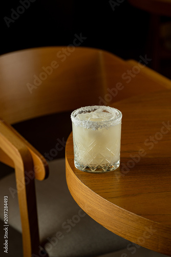 A drink sits on a wooden table next to a comfortable chair. The drink has ice and a salt rim. The environment is dimly lit, creating a warm feeling at night