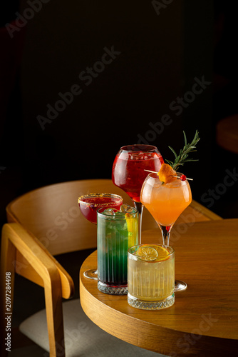 Four drinks with different colors sit on a wooden table in a dimly lit bar. The setting is lively, and the drinks are ready to be enjoyed by guests
