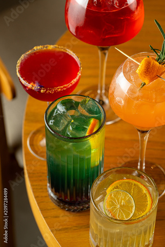 Five different cocktails are placed on a wooden table. Each drink has unique colors and garnishes. Friends may enjoy these drinks together in a lively atmosphere