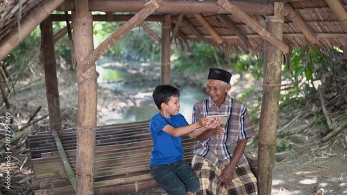 Young boy stands beside smiling elderly man beneath rustic bamboo shelter, both focused on smartphone in shared hands, surrounded by woven roof, wooden platform, calm water, and soft daylight