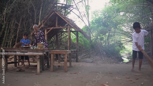 Elderly woman and two children stand beside rustic shelter in tropical village clearing, while one child sweeps ground near simple wooden tables amid dense greenery and soft daylight