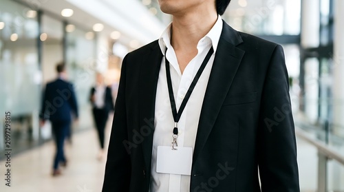 Young male professional wearing blank ID badge on lanyard in bright modern office space with blurred colleagues walking in busy corporate work environment