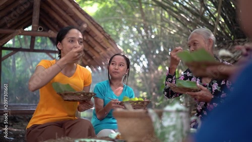 Three generations sit together beneath timber shelter, eating from woven baskets lined with leaves while sharing relaxed conversation in warm daylight amid tropical surroundings, with family, eating