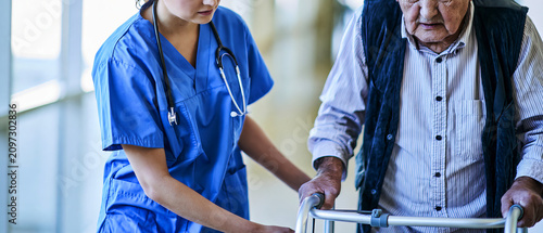  young female nurse  assisting a senior  man using a walker in a brightly lit hospital corridor