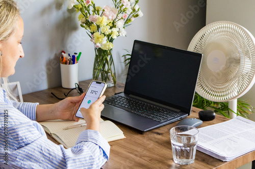 Woman sitting at home office desk holding smartphone displaying indoor temperature of 32 degrees Celsius beside laptop. Concept of monitoring indoor climate, heatwave awareness, smart home technology