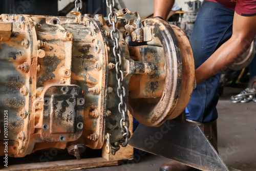 Foto Close up of mechanic hands working on a rusty heavy truck transmission gearbox suspended by a metal chain hoist in a garage