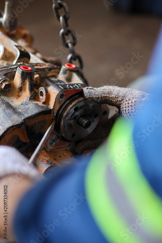 Fotografie Close-up of a mechanic in protective gloves using a flat screwdriver to pry and disassemble a heavy metallic truck gearbox suspended on a hoist chain