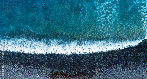 Top down aerial view with abstract background of blue turquoise sea waves crashing on a black pebble beach with white foam on a dark volcanic stone shore