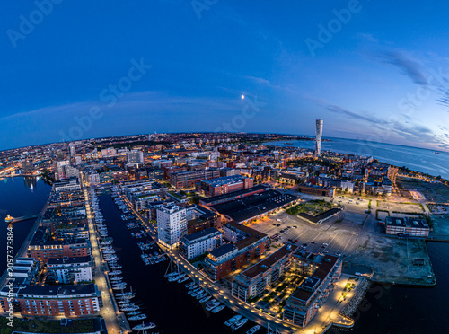 Aerial drone shot of Malmö harbor in twilight, blue hour glow, summer evening 2024, with city lights, reflections, and Scandinavian urban landscape beauty.