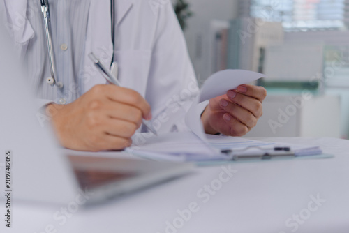 Hands of a physician in a white lab coat writing on clinical documents with a pen. The medical professional signs a prescription or patient report on a clipboard at a bright office workspace.